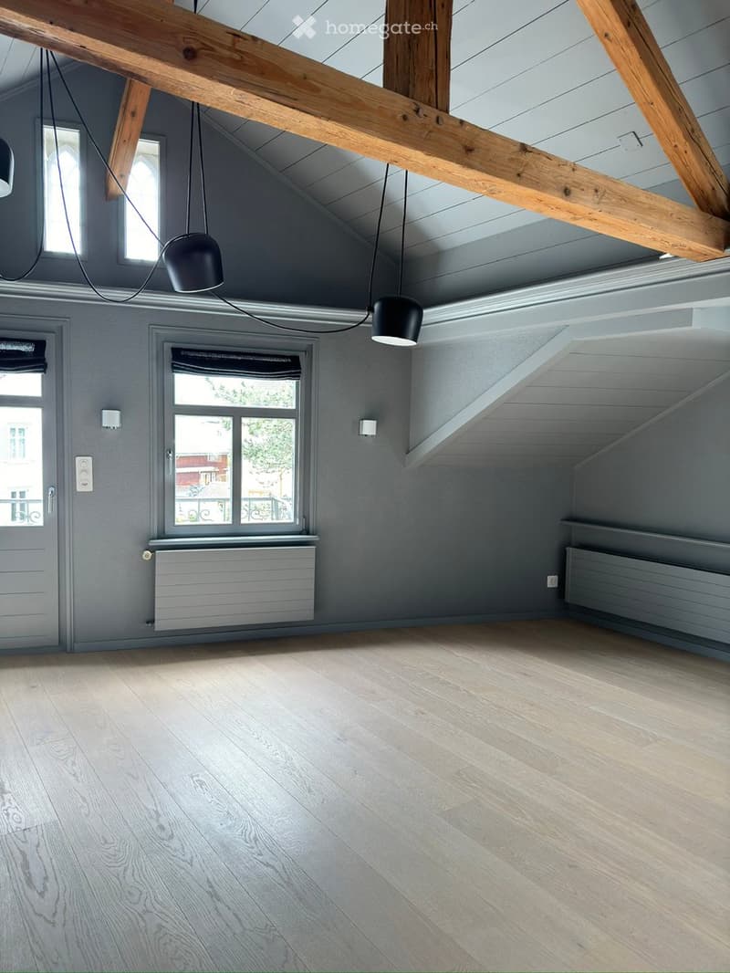 Empty attic room with wooden beams, exposed ceiling, wooden flooring, and large windows. No furniture or equipment present.