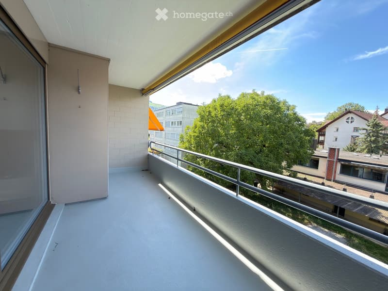 Balcony with white flooring, metal railing, and large windows offering a view of trees and buildings.