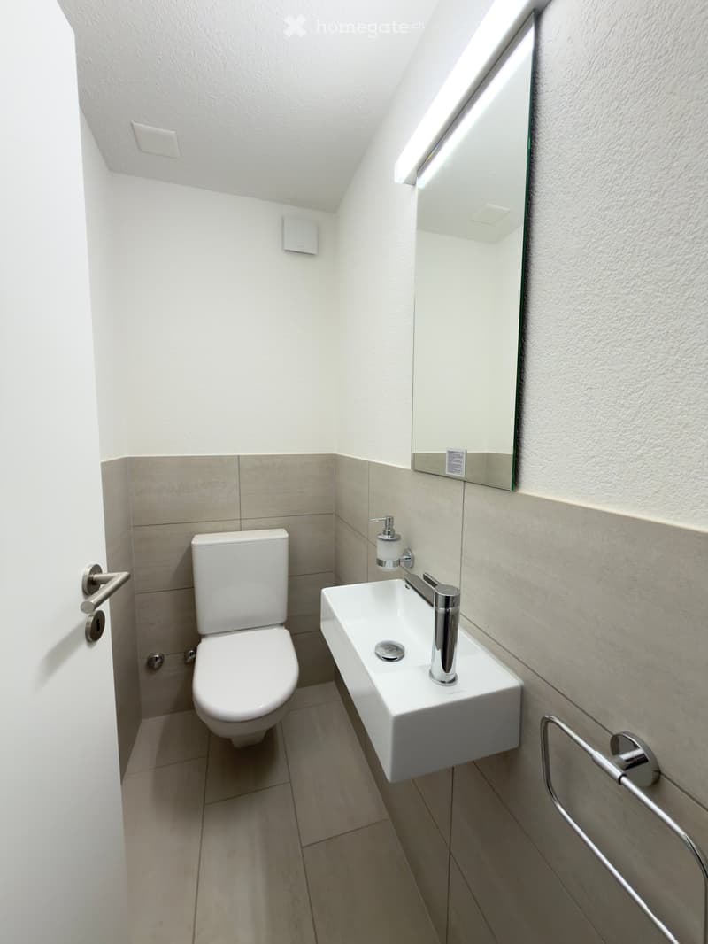 Modern, compact bathroom with white tiles, a toilet, a wall-mounted sink, a mirror, and a soap dispenser.