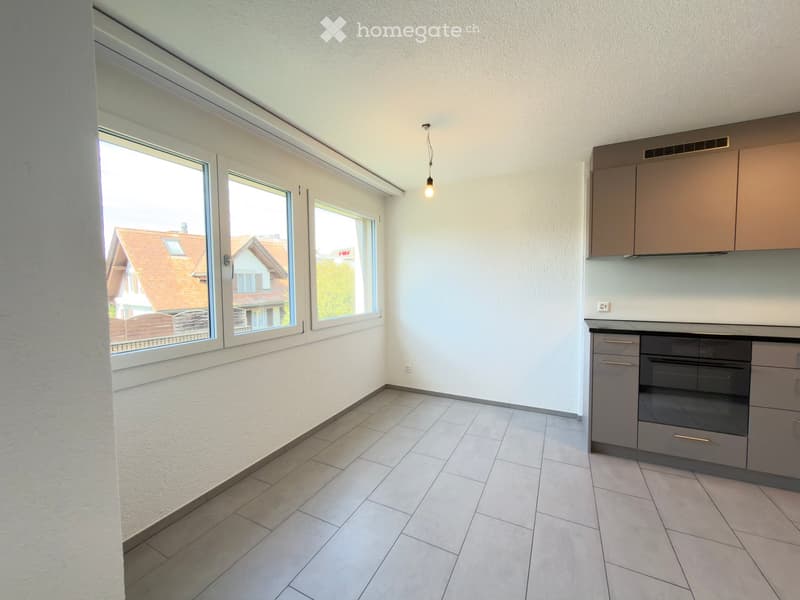 Empty kitchen with tiled floor, light-colored walls, and built-in cabinets. Large windows offer a view of neighboring houses.