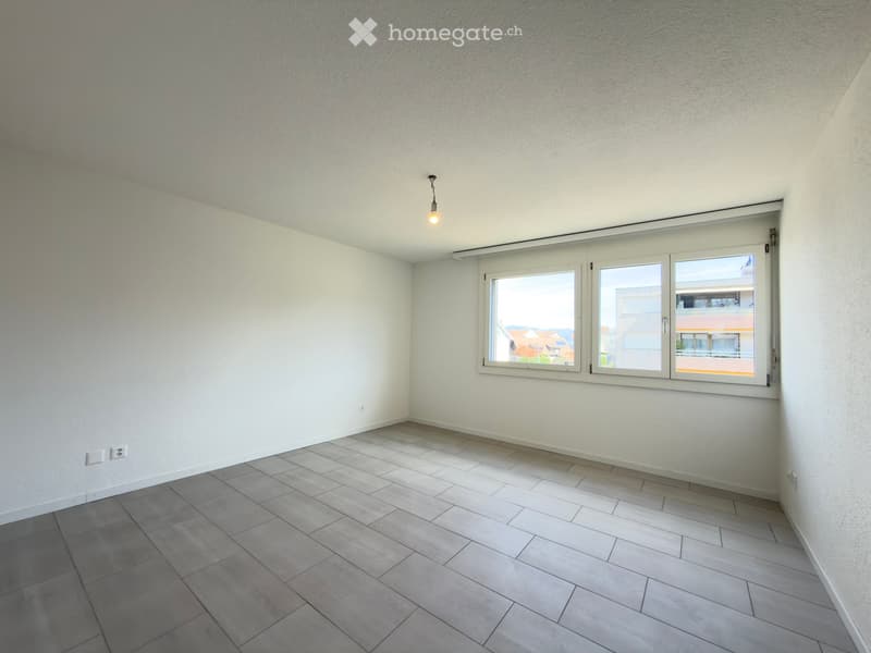 Empty room with light-colored walls, tiled floor, and large windows offering a view of the outside area.