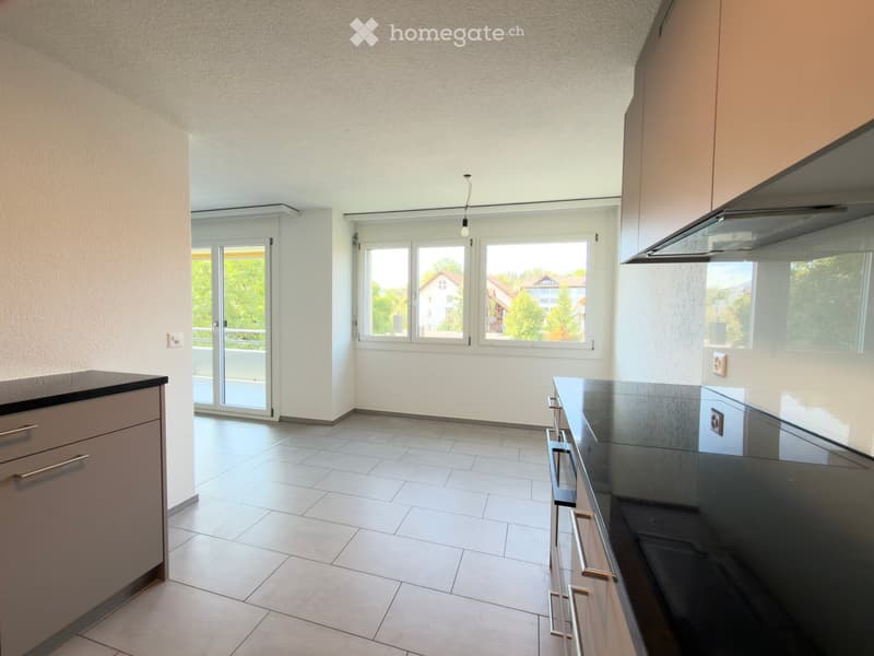 Empty kitchen with white tiled floor, light-colored cabinets, and large windows overlooking a green area.