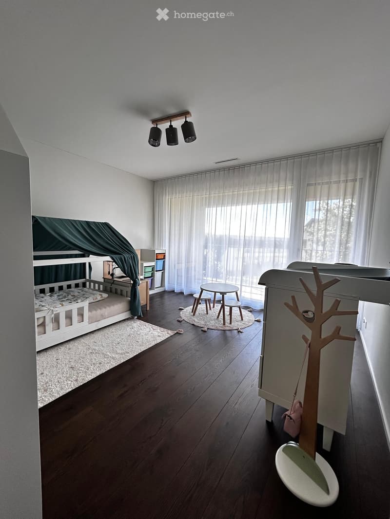 A well-lit children's bedroom with a bunk bed, a small round table with stools, a storage unit, and a decorative tree-shaped toy. The room features wooden flooring, large windows with sheer curtains, and modern lighting.
