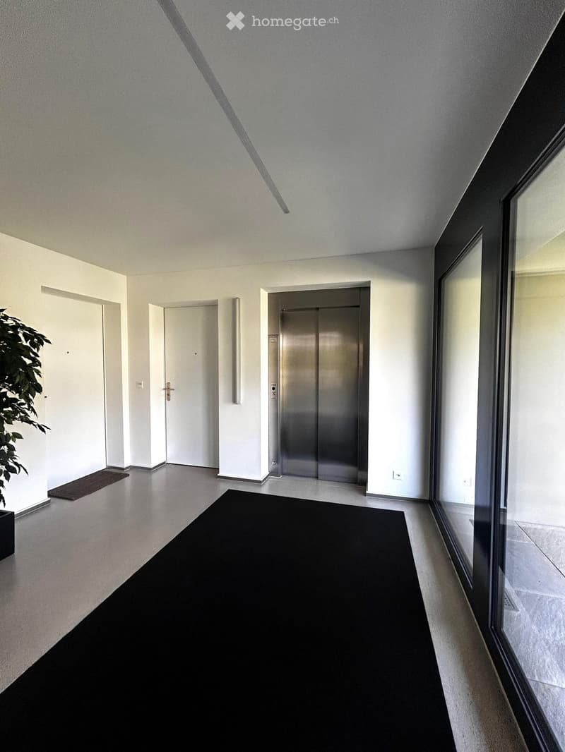 Modern hallway with white walls, polished floor, elevator, and large windows on one side.