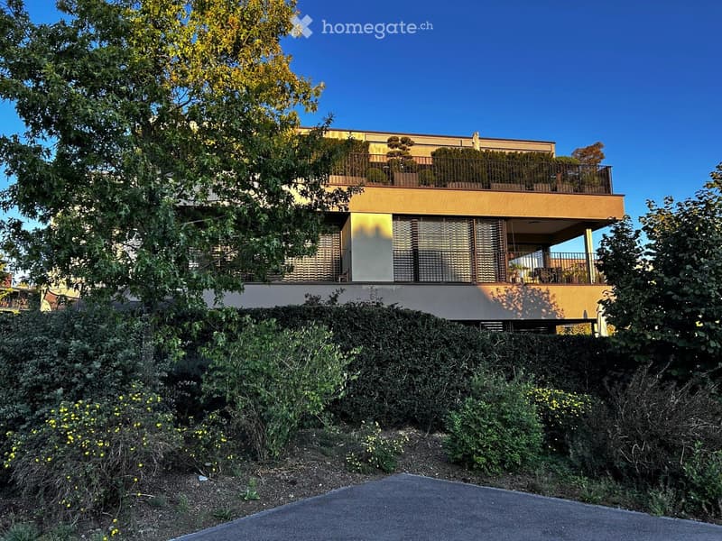 Modern two-story house with large windows, balconies, and surrounding greenery.