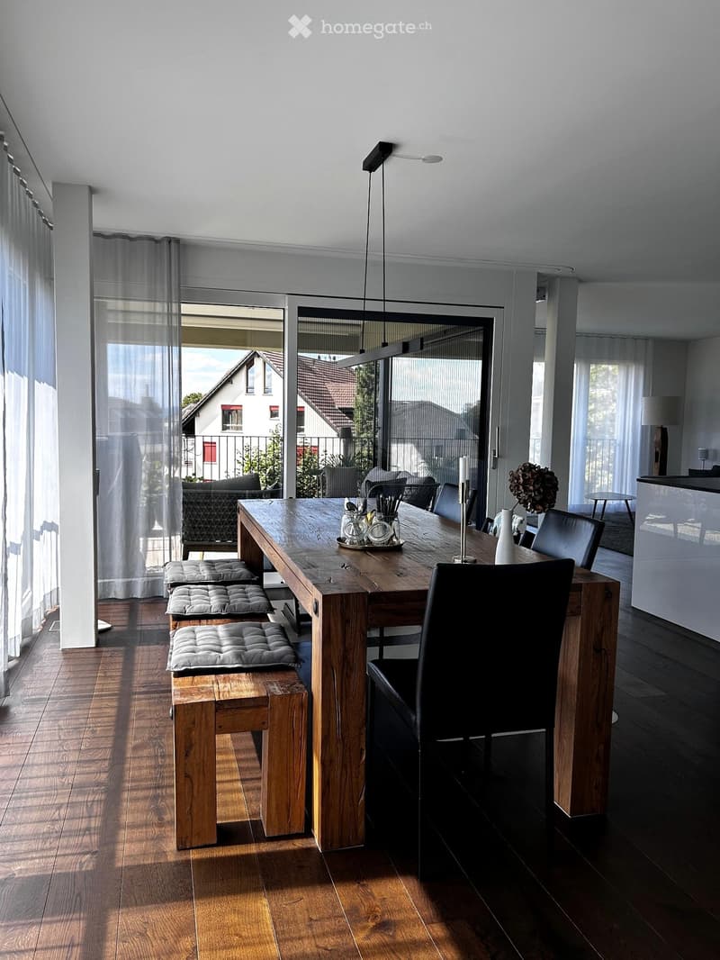 Bright, modern kitchen with a large wooden dining table, black chairs, wooden stools, and a view of the outdoor area through large windows.
