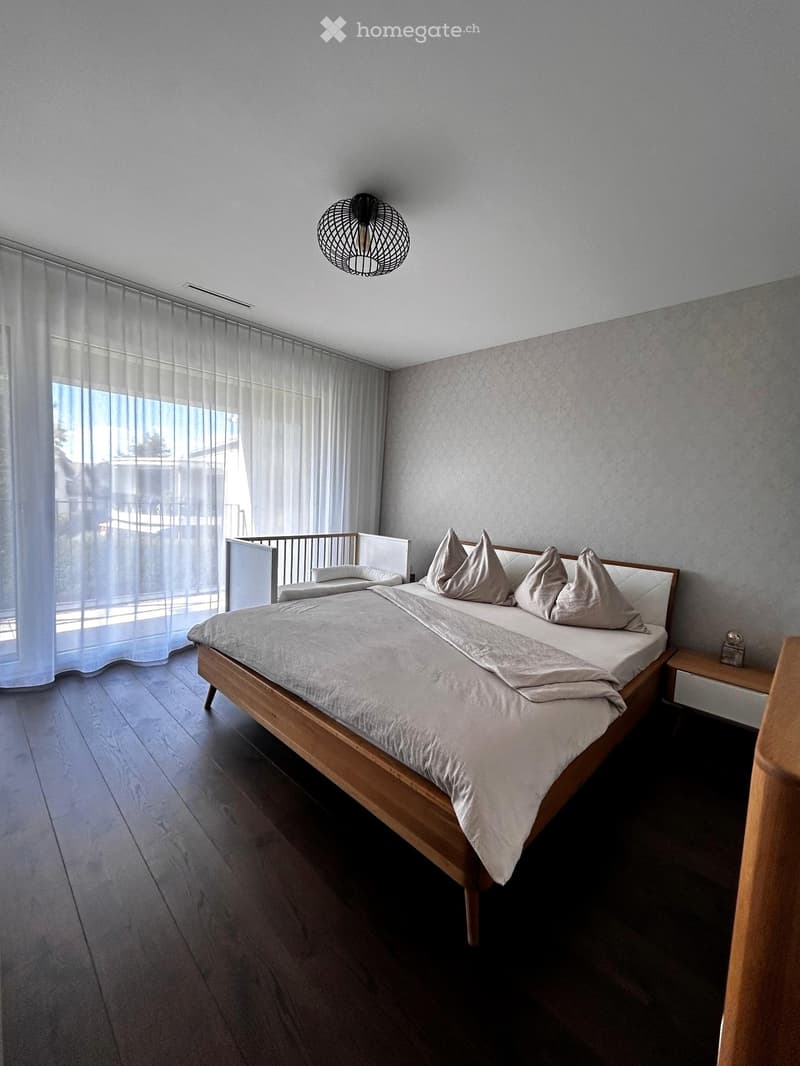 Modern bedroom with a wooden bed, light-colored bedding, and a pendant light fixture. Large windows with sheer curtains allow natural light to enter.
