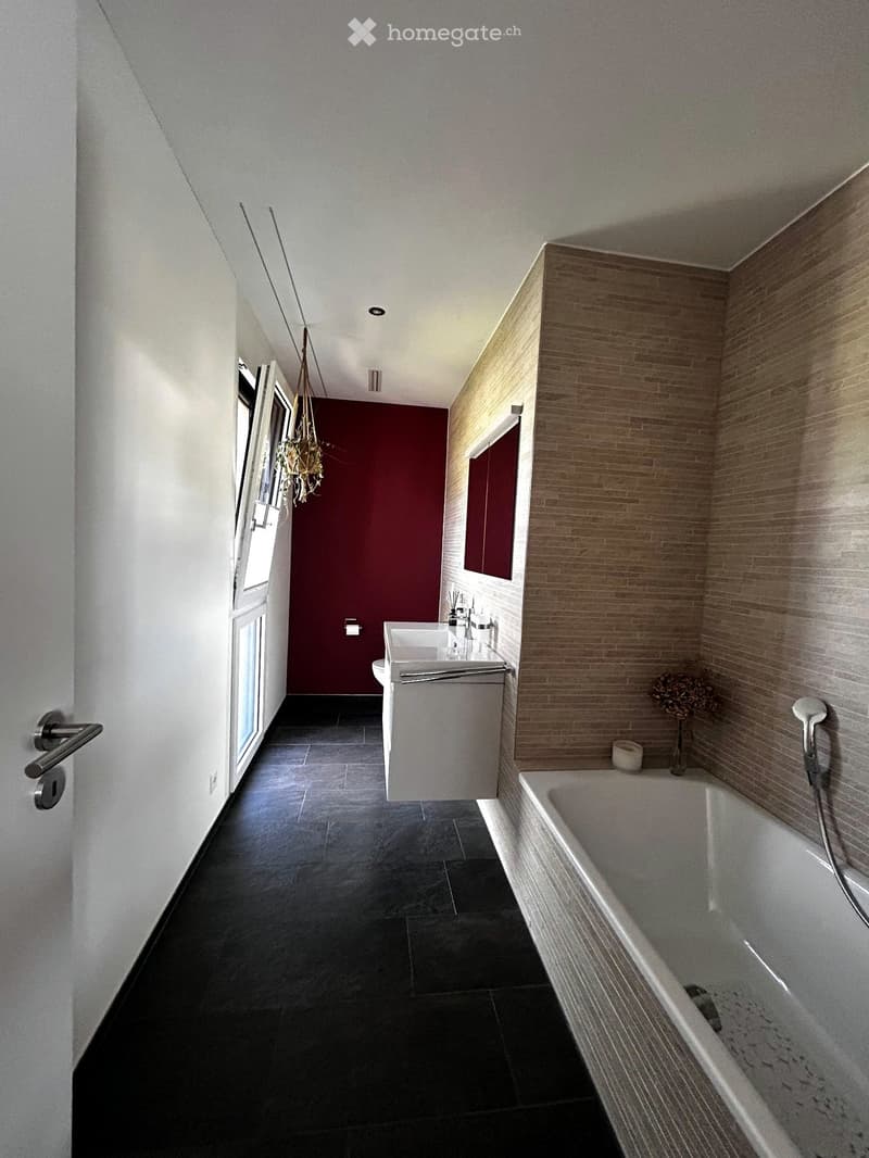 Modern bathroom with dark tiled floor, white bathtub, sink, and mirror. Features a red accent wall and a hanging decorative item.
