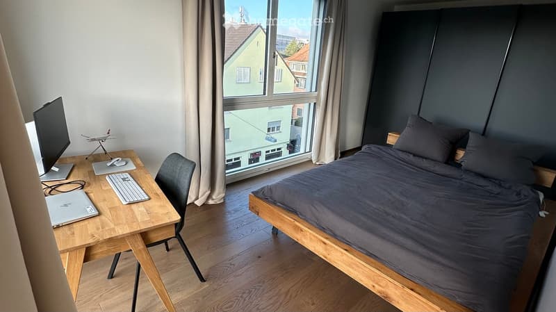 Modern bedroom with a wooden bed, desk with laptop, keyboard, mouse, and screen, and a model airplane on the desk. Large window with a view of the city.
