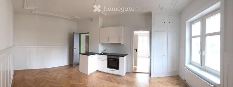 Bright, unfurnished room with white walls, wooden flooring, and built-in white cabinets. Features a small kitchen area with a sink, oven, and countertop.