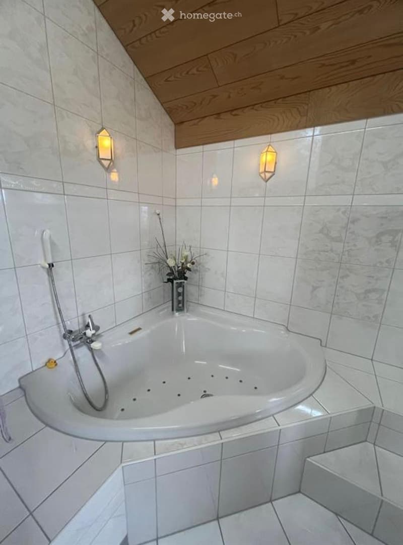 Modern bathroom with a large, oval bathtub, tiled walls, and decorative wall-mounted lights. A vase with flowers sits on the edge of the tub.