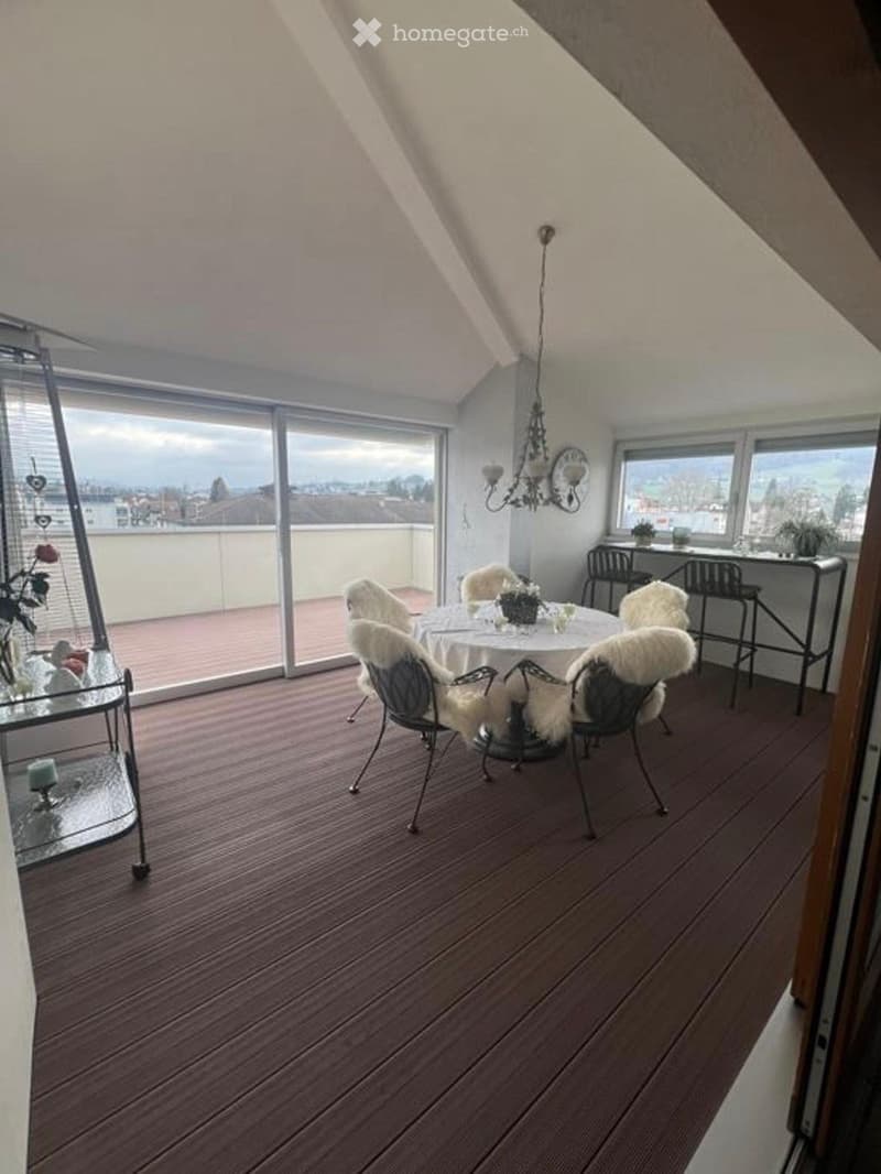 Spacious balcony with wooden flooring, large windows offering a scenic view, a round dining table with chairs, and a chandelier.