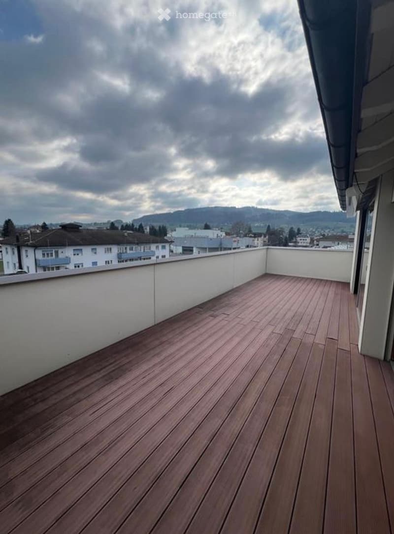Wooden deck with white railings, overlooking a cityscape with buildings and hills under a cloudy sky.