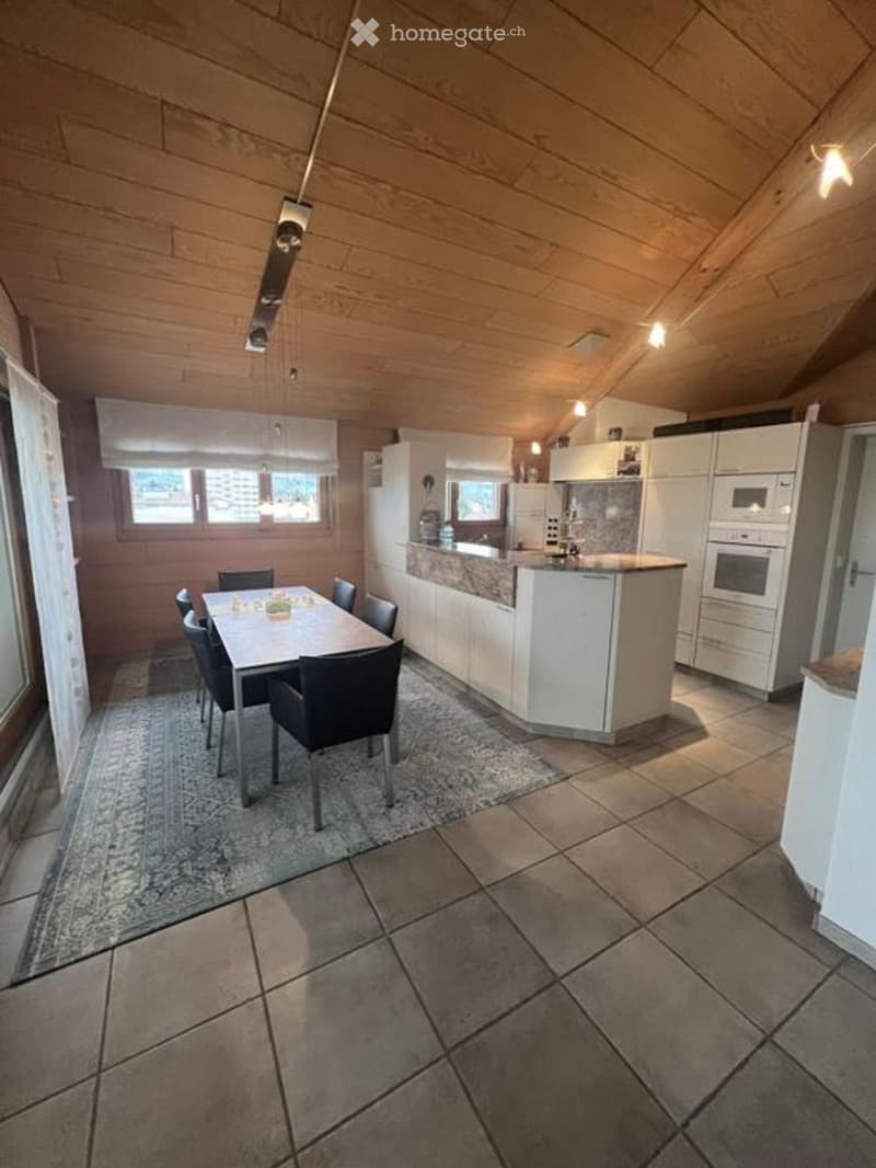 Open-plan kitchen and dining area with white cabinets, a central island, a dining table with chairs, and modern appliances including a refrigerator, oven, and microwave. The space features wooden ceilings and tiled flooring.