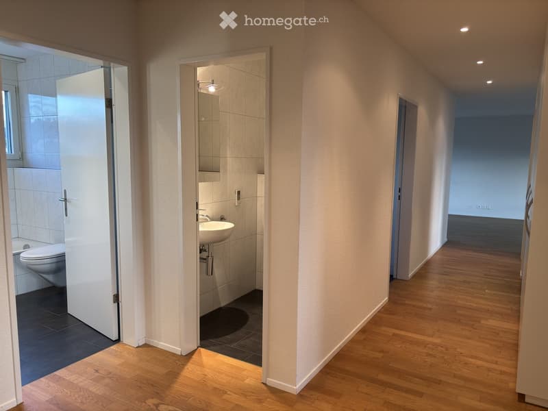 A well-lit hallway with wooden flooring, leading to a bathroom and another room. The bathroom features a sink, toilet, and tiled walls.