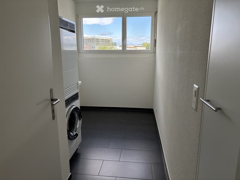 Empty room with tiled floor, grey walls, and a washing machine in the corner. Large window with a view of the outside.