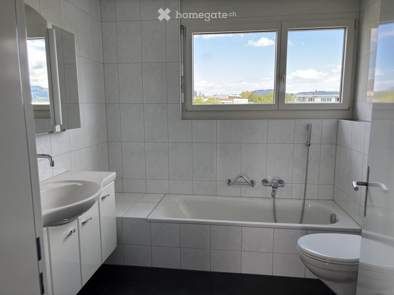 Modern bathroom with white tiles, a bathtub, sink, toilet, and a large window offering a scenic view.