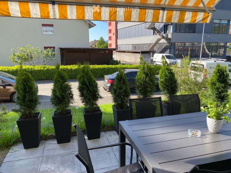 Balcony with a table, chairs, and potted plants, surrounded by greenery and neighboring buildings.