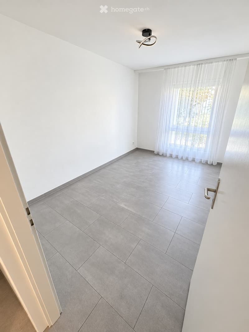Empty room with white walls, grey tiled floor, large window with sheer curtains, and a modern ceiling light fixture.