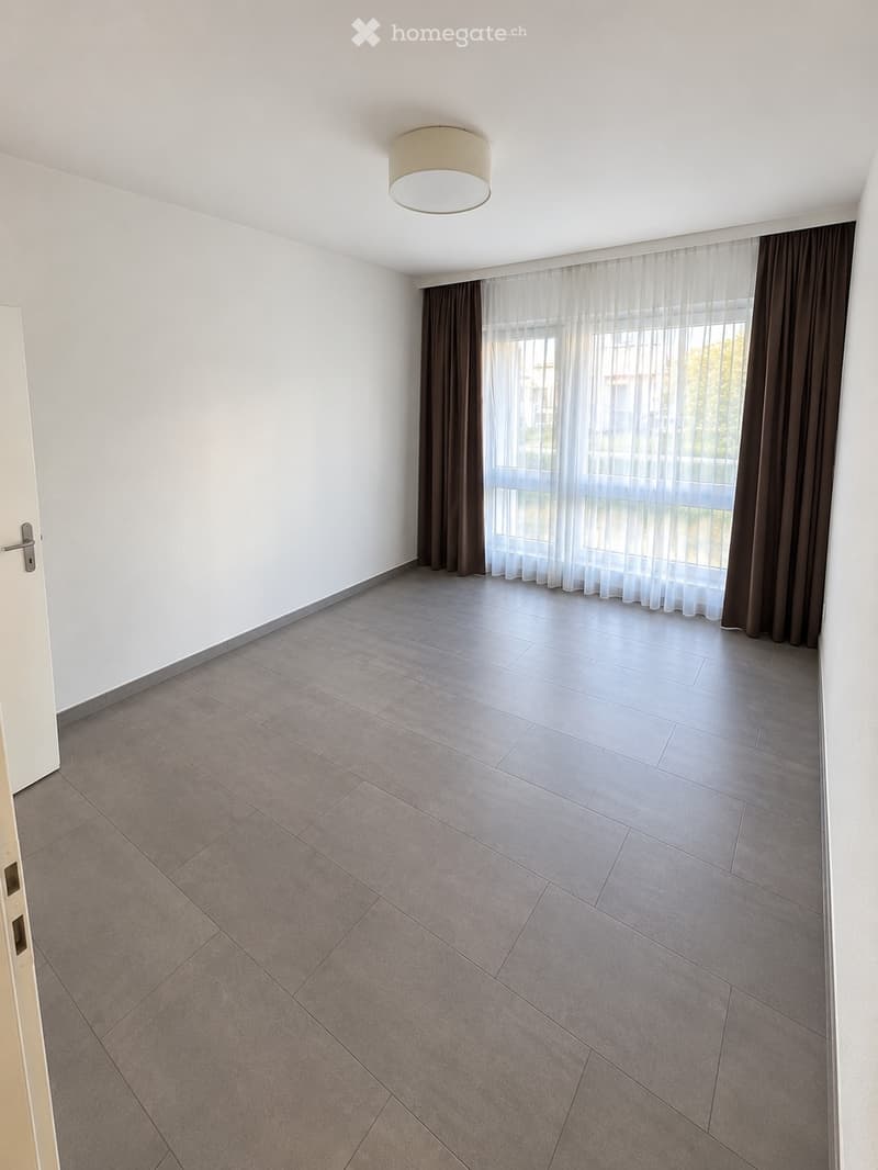 Empty room with grey tiled floor, white walls, large window with sheer curtains and dark drapes, and a round ceiling light fixture.