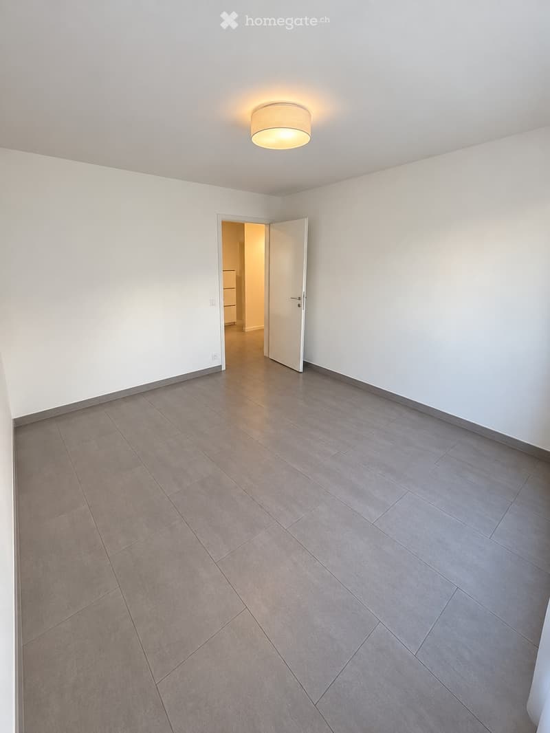 Empty room with white walls, grey tiled floor, and a single ceiling light fixture. An open door leads to another room.
