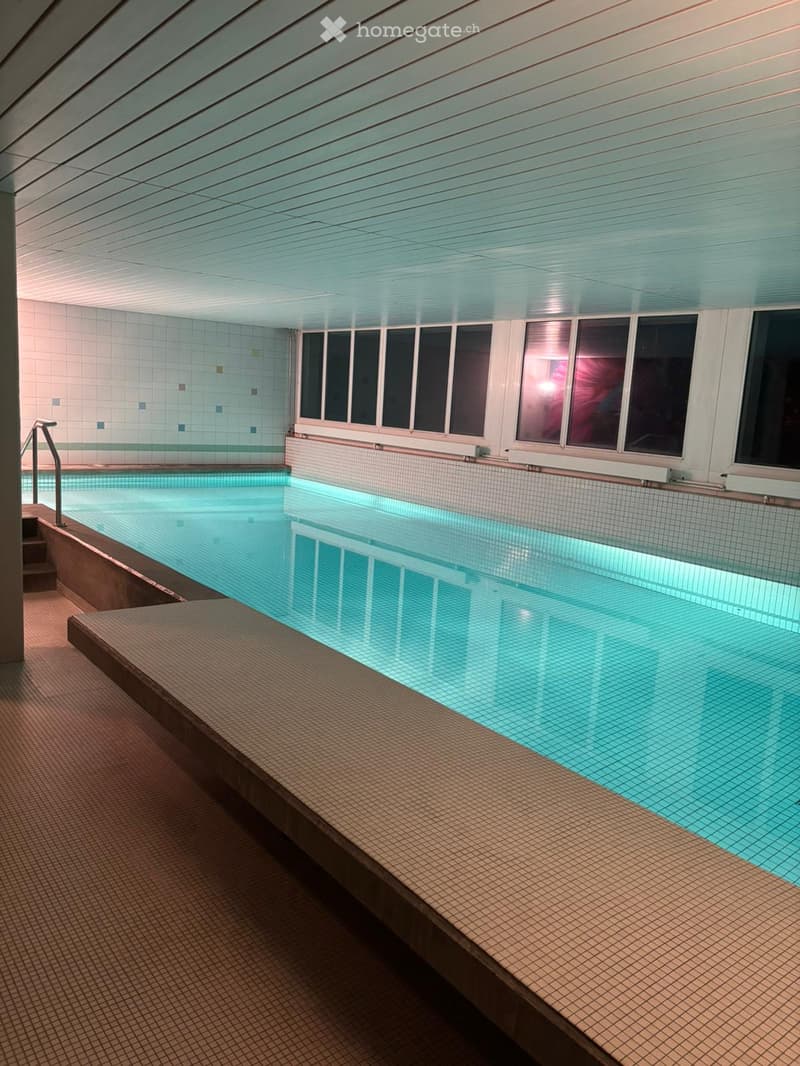 Indoor swimming pool with clear water, tiled walls, and a staircase leading into the pool. The ceiling has a modern design with recessed lighting.