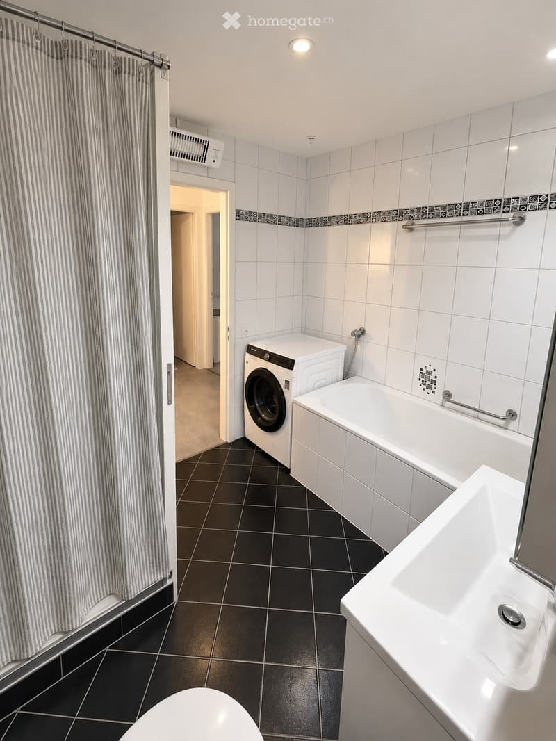 Modern bathroom with white tiles, black floor tiles, bathtub, sink, toilet, washing machine, and shower curtain.