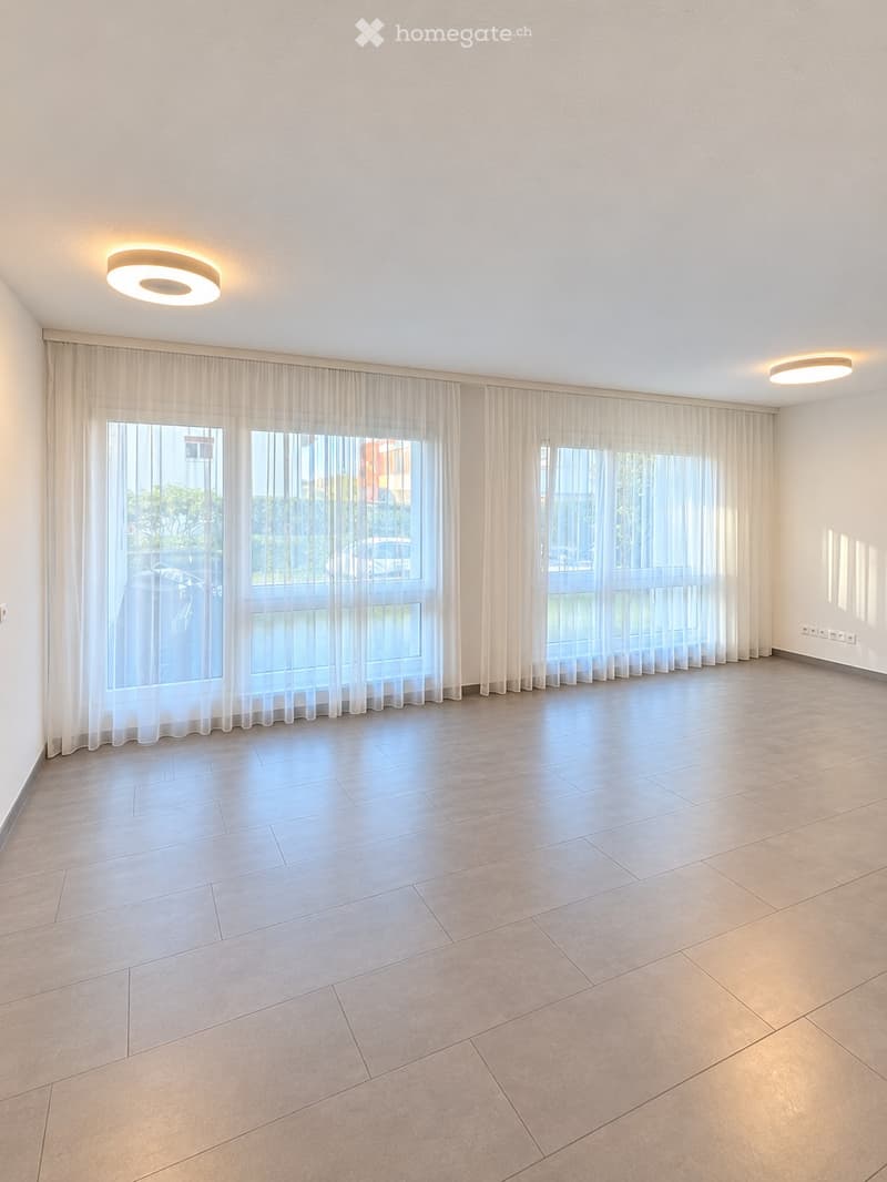 Empty room with tiled floor, large windows with sheer curtains, and recessed ceiling lights.