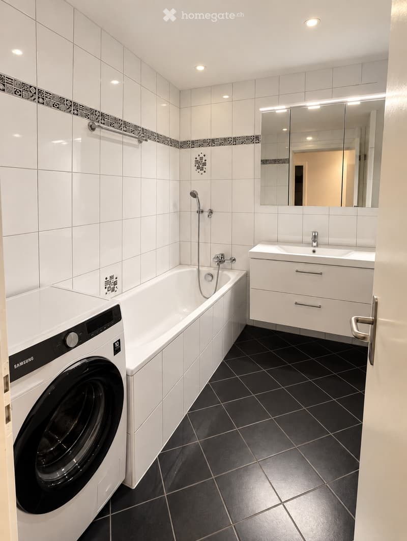 Modern bathroom with white tiles, black tiled floor, bathtub, sink, mirror, and a Samsung washing machine.