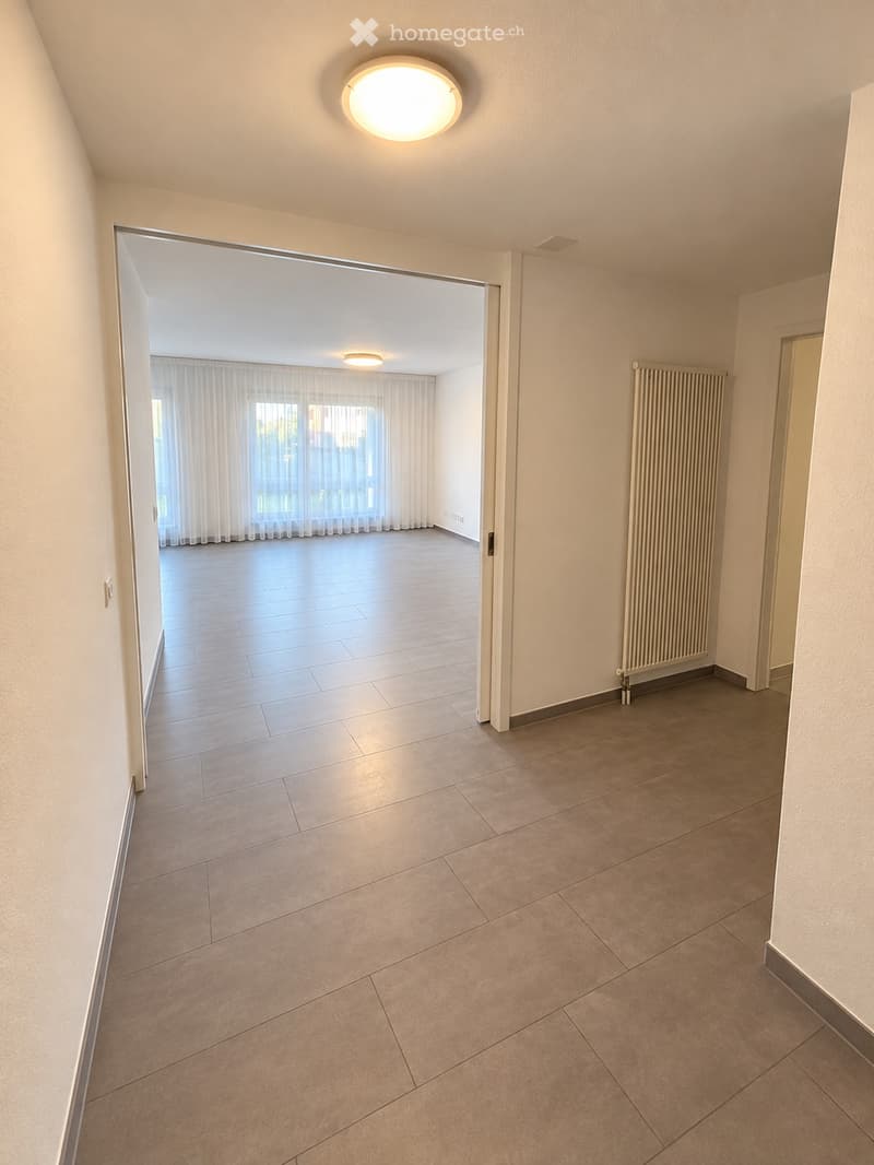 Empty room with tiled floor, white walls, and a radiator. An open doorway leads to another room with large windows and white curtains.
