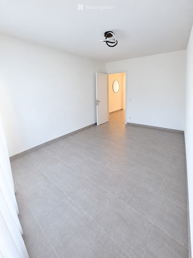 Empty room with grey tiled floor, white walls, and a modern ceiling light fixture. An open doorway leads to another room with a visible mirror on the wall.