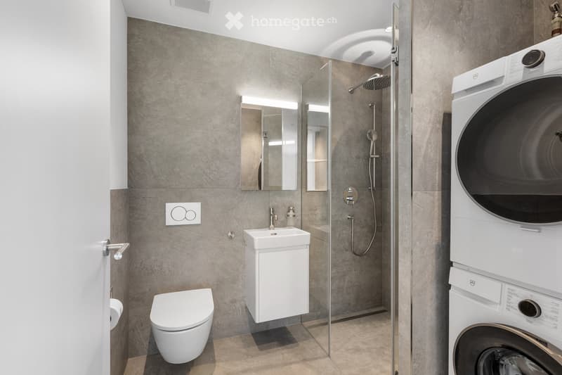 Modern bathroom with a toilet, sink, mirror, glass-enclosed shower, and stacked washing machine and dryer.