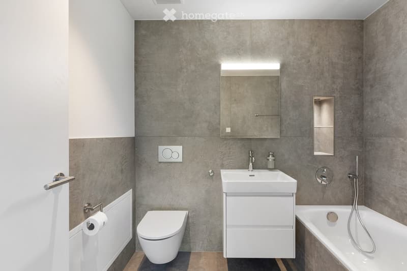 Modern bathroom with grey tiled walls, white fixtures including a toilet, sink, bathtub, and mirrored cabinet.