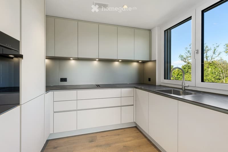 Modern kitchen with white cabinets, stainless steel sink, and a sleek faucet. The space features a large window with a view of greenery outside.