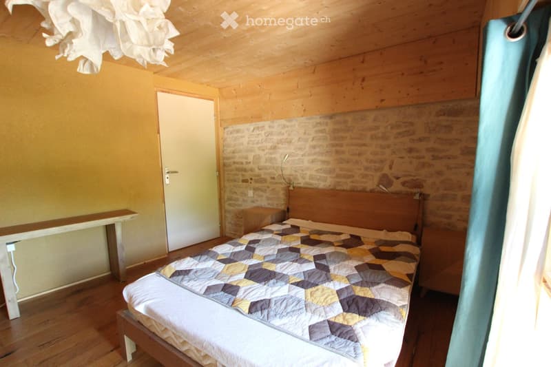Cozy bedroom with wooden bed, quilted bedspread, nightstand, and bench near the door. Exposed wooden ceiling and brick accent wall.