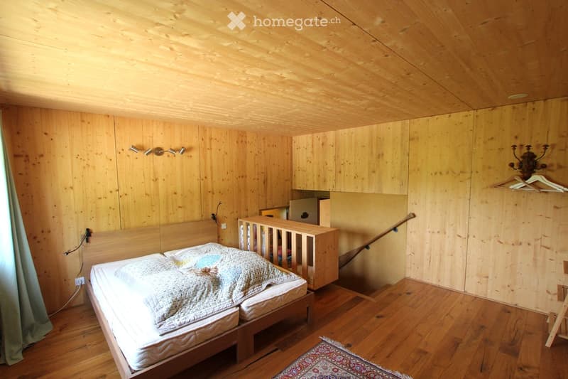 Cozy wooden bedroom with a double bed, a small cradle, built-in wardrobe, and a mirror. The room features wooden walls and ceiling, a small rug on the floor, and warm lighting.