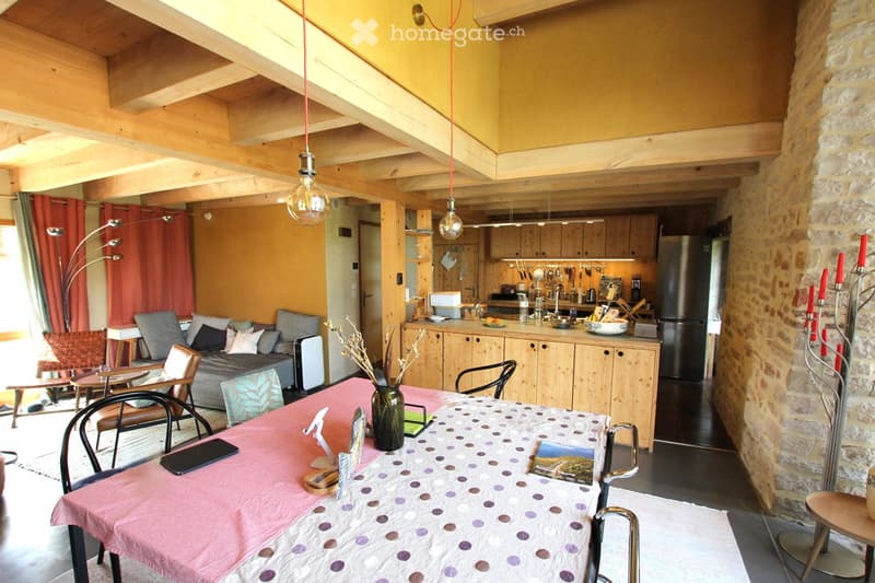 A cozy, well-furnished living and dining area with exposed wooden beams, a sofa, dining table, chairs, and a kitchen area with cabinets and appliances.