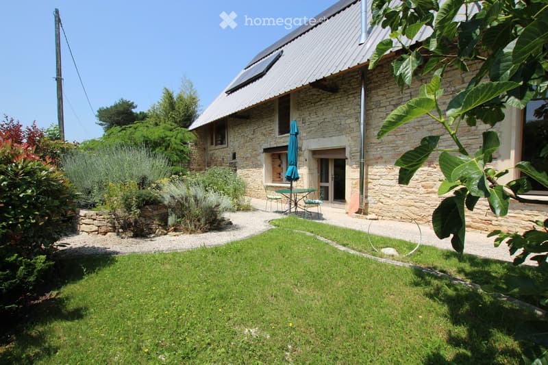 Stone house with a metal roof, surrounded by a well-maintained garden with a gravel pathway, green lawn, and outdoor seating area with a table and chairs.