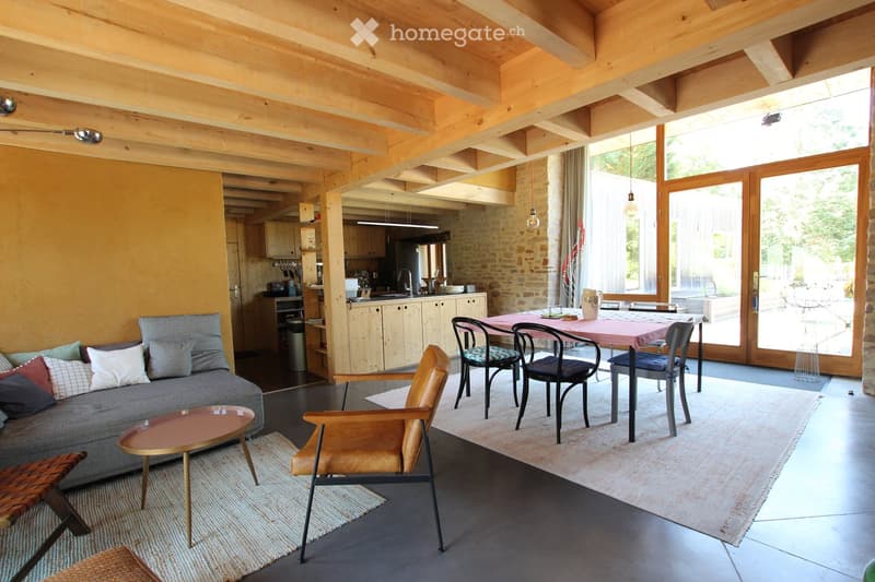 Open-plan living and dining area with exposed wooden beams, grey sofa, coffee table, dining table with chairs, and a modern kitchen area with cabinets and appliances.