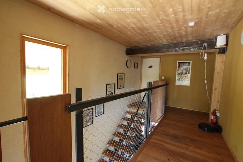 Hallway with wooden flooring, exposed wooden ceiling beams, and a staircase with a metal railing. Several framed pictures and posters are displayed on the walls.