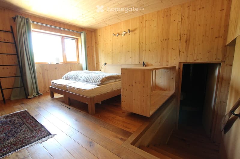 Cozy wooden bedroom with a double bed, nightstand, and a small rug on the floor. Natural light streams in from a window with wooden frames and curtains.