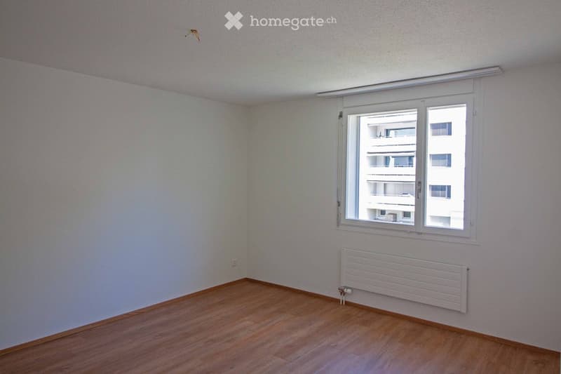 Empty room with light-colored walls, wooden flooring, a window with white frame, and a white radiator below the window.