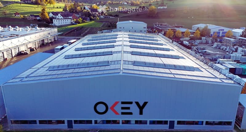 Aerial view of a large industrial building with a modern design, featuring solar panels on the roof and the logo "OKEY" prominently displayed on the facade. The surrounding area includes other industrial structures, parking lots, and open fields.
