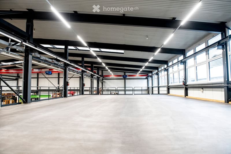 Large, open industrial-style space with a concrete floor, exposed metal beams, and large windows allowing natural light. Minimal furnishings, with a few storage units and a small staircase visible in the background.