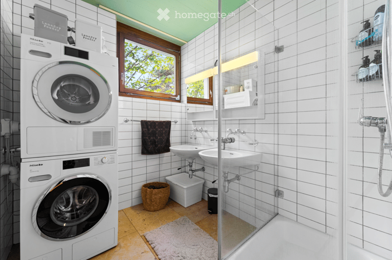 Modern bathroom with white tiled walls, a washing machine, two sinks, a shower, and a window with a view of greenery.