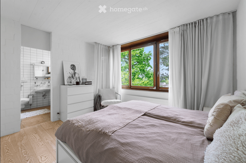 Bright bedroom with wooden flooring, large window with view, bed with pillows and blanket, white dresser, chair, and adjacent bathroom with sink and toilet.
