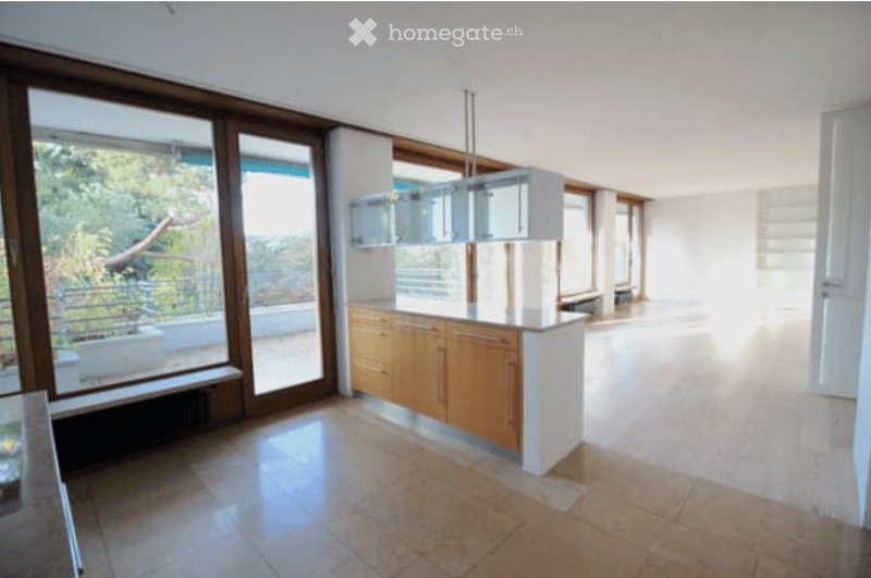 Bright, open-plan kitchen and living area with large glass doors leading to a balcony. The space features modern wooden cabinets, a white countertop, and a sleek pendant light fixture.