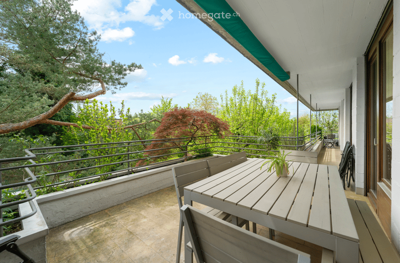 Balcony with a wooden table, chairs, and a potted plant, overlooking a lush green garden with trees and a clear blue sky.