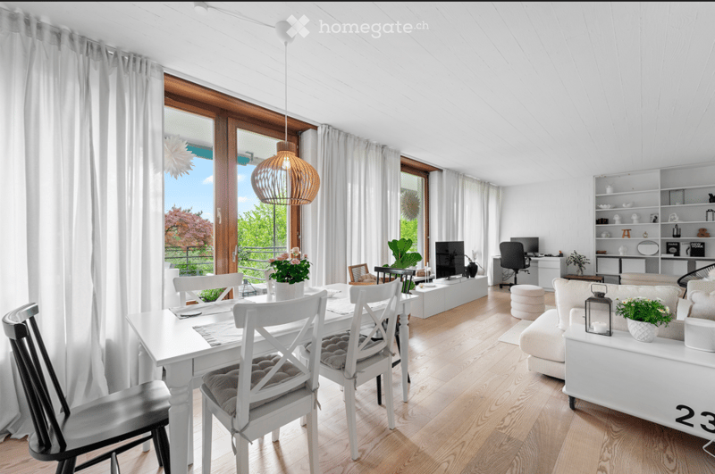 Bright, modern living and dining area with white furniture, large windows, and a balcony view. Features include a white sofa, coffee table, dining table with chairs, TV, shelves, and decorative plants.