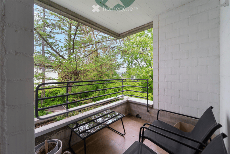 Balcony with white brick wall, metal railing, outdoor furniture including a table and chairs, overlooking a green, tree-filled area.